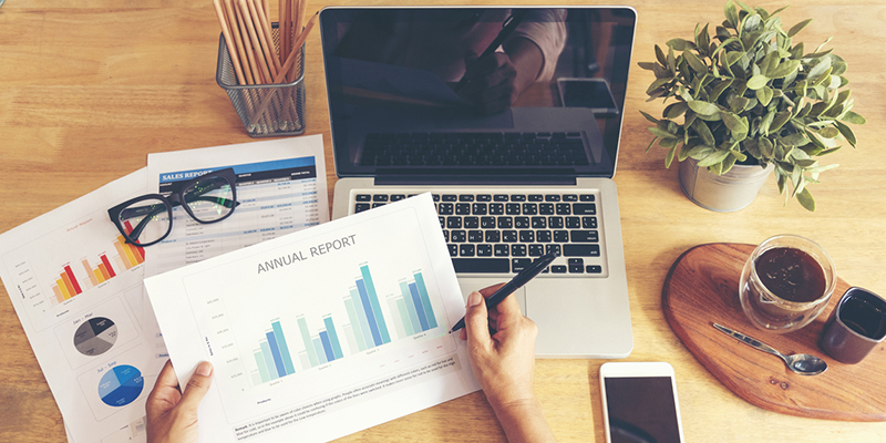 Woman working at home office desk using laptop business financial document chart and graph on wooden table with coffee cup. Woman freelance reading business graphs sale report on business office desk
